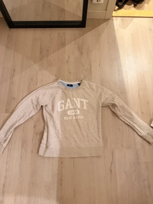 Beige sweatshirt från GANT - Säljer en beige sweatshirt från GANT med tryck 'GANT 1949 NEW HAVEN' på bröstet. Tröjan har rund halsringning, långa ärmar och ribbade muddar. Perfekt för dig som gillar klassisk och avslappnad stil.