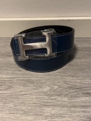 H-bälte  - Snyggt bälte i blått och svart skinn med ikoniskt,H-spänne i silverfärgad metall. Bältet har stilrena, kontrastsömmar och klassisk design som lyfter, vilken outfit som helst. Strl 110cm