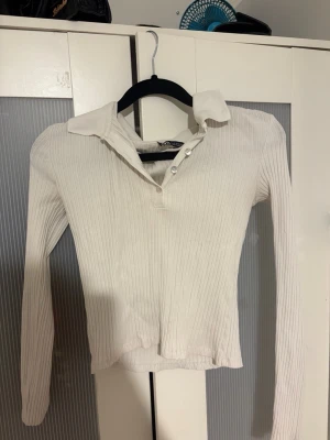 Ribbad vit långärmad polotopp - Säljer en ribbad vit långärmad polotopp med krage och knappar framtill. Toppen har en smickrande passform och är tillverkad i mjukt material som känns skönt mot huden. Perfekt att styla med jeans eller kjol för en clean look.