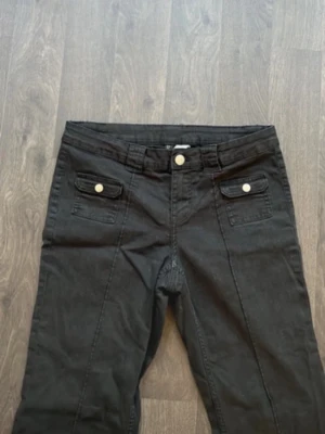 Svarta bootcut byxor med fickdetaljer - Säljer ett par svarta bootcut byxor med snygga fickor framtill som har lock och silverfärgade knappar. Byxorna har normal passform och är tillverkade i ett mjukt bomullsmaterial. Perfekta till en chill vardagslook.