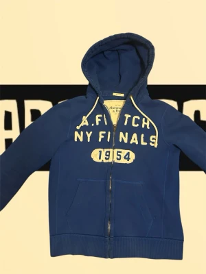 Blå zip hoodie från Abercombie & Fitch  - Stilren och grisch blå huvtröja från Abercrombie & Fitch, storlek M. Tillverkad i mjukt bomullsmaterial, perfekt för vardagsbruk. Tröjan har dragkedja fram, kängurufickor och broderad text på bröstet. Använd men i gott skick, lite slitage på trycket vilket ger en snygg vintagekänsla. Normal passform, passar bra till både jeans och träningsbyxor.