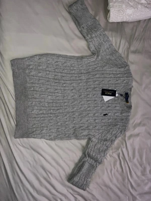 Grå stickad tröja Polo Ralph Lauren - Säljer en snygg grå kabelstickad tröja från Polo Ralph Lauren i storlek M. Tröjan har rund hals, långärmade ribbade muddar och klassisk broderad logga på bröstet. Perfekt för dig som gillar stilrena och tidlösa plagg med premiumkänsla. Obs kommer ej med tagg!