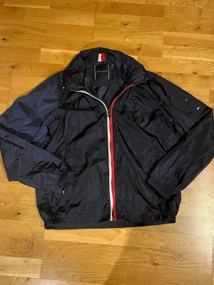 Marinblå vindjacka från Tommy Hilfiger - Snygg marinblå vindjacka från Tommy Hilfiger med klassiska röda och vita detaljer längs dragkedjan. Jackan har lång ärm, elastiska muddar och en ficka med knapp på ärmen. Tillverkad i polyester och perfekt för vår och höst.