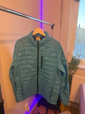 Ljusblå McKinley pufferjacka - Säljer en ljusblå pufferjacka från McKinley med quiltad design och hel dragkedja framtill. Jackan har en bröstficka med dragkedja, två sidofickor och en hög krage. Perfekt för kyliga dagar och enkel att matcha med olika outfits.