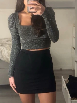 Glittrig croppad topp med puffärm - Säljer en glittrig mörkgrå croppad topp med långa puffärmar och fyrkantig ringning. Toppen har en tajt passform och skimrar snyggt i ljuset, perfekt för att sticka ut. Materialet känns mjukt och stretchigt.