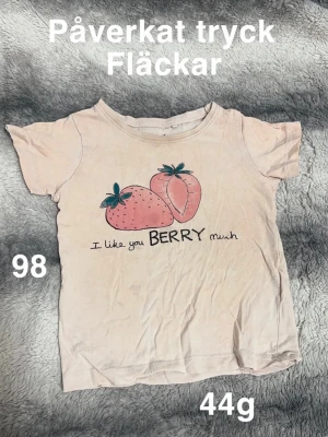 Ljusrosa t-shirt med jordgubbar - Söt ljusrosa t-shirt med tryck av två jordgubbar och texten 'I like you BERRY much' framtill. Klassisk rund hals och korta ärmar. Perfekt för dig som gillar gulliga motiv och mjuka färger.🌸 Påverkat tryck. Fläckar. Storlek 98