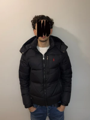 Svart pufferjacka från Ralph Lauren - Säljer en svart pufferjacka från Ralph Lauren med huva och dragkedja framtill. Jackan har en röd broderad logga på bröstet och är vadderad för extra värme. Perfekt för kyliga dagar och har en modern, stilren look.