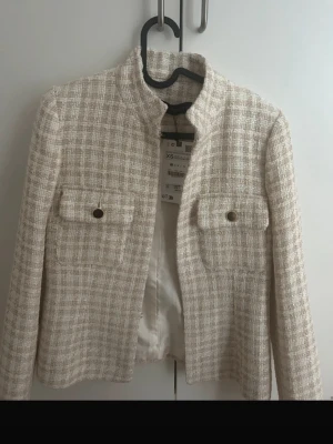 Beige rutig kavaj från Zara XS - Säljer en beige rutig kavaj från Zara i storlek XS. Jackan har två fickor framtill med guldknappar och en rak, klassisk passform. Materialet är strukturerat och ger en lyxig känsla. Perfekt för att lyfta vilken outfit som helst.