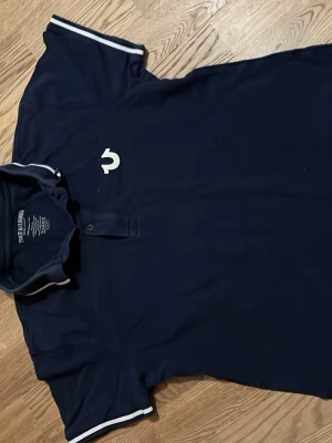 Marinblå pikétröja True Religion S - Säljer en marinblå pikétröja från True Religion i storlek S. Tröjan har vit logga på bröstet och stort tryck på ryggen. Klassisk krage med knappar och vita ränder på ärmslut och krage. Tillverkad i 100% bomull för en skön känsla.