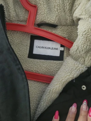 Svart parkas från Calvin Klein Jeans - Säljer en svart parkas från Calvin Klein Jeans med mysigt teddyfoder och huva med fuskpäls. Jackan har fyra stora fickor framtill och stängs med dragkedja och knappar. Perfekt för kalla dagar och riktigt snygg streetstil.