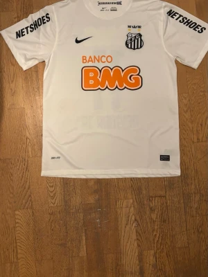 Vit Santos andratröja Neymar JR 11 - Santos FC matchtröja i vitt med svarta detaljer, Neymar JR och nummer 11 på ryggen. Tröjan har tryckta sponsorer som BMG och Netshoes, samt klubbmärke och Nike-logga på bröstet. Tillverkad med DRI-FIT teknologi