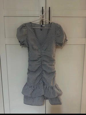 Svartvit rutig kortklänning med volanger - Supersöt kortklänning från H&M i svartvitt rutigt mönster. Puffärmar, v-ringning och rynkade detaljer framtill. Volanger nertill ger extra flow och klänningen har en figurnära passform. Perfekt för dig som gillar en lekfull och trendig stil.