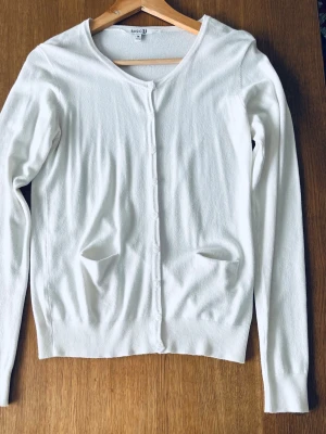 Vit kofta från BASICU med knappar - Säljer en stilren vit kofta från BASICU i storlek M. Koftan har rund hals, långa ärmar och knäppning framtill med små knappar. Tillverkad i mjuk viskosblandning med två fickor framtill. Perfekt att slänga över en topp för en clean look.