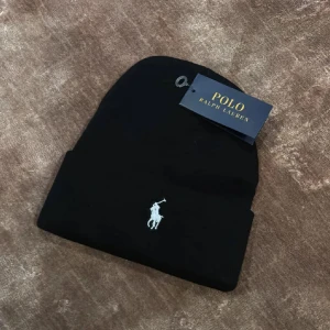 Svart mössa från Polo Ralph Lauren - Klassisk svart mössa från Polo Ralph Lauren med den ikoniska broderade loggan framtill. Mössan är ribbstickad och har en vikt kant för extra stilpoäng. Perfekt accessoar för kyliga dagar.
