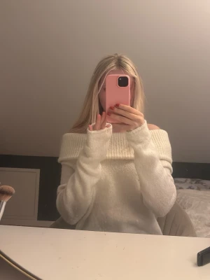 Vit offshoulder stickad tröja - Mysig vit stickad tröja med bred ribbad kant och offshoulder-design. Tröjan har lång ärm och en loose passform som ger en avslappnad och trendig look. Perfekt för kyliga dagar när du vill vara både bekväm och snygg.