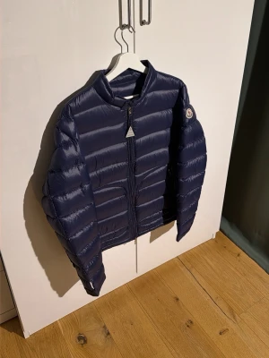 Mörkblå Moncler pufferjacka - Säljer en mörkblå pufferjacka från Moncler med klassisk logga på ärmen. Jackan har quiltad design, dragkedja framtill och hög krage. Tillverkad i glansig polyester och dunfylld för extra värme. Size 3. Defekt på bild 3! Ihop klistrad med en tygklisterlapp, funkar utmärkt! Toppen i skick overall🤗Endast defekten som försämrar skicket lite annars som en ny jacka 🧥 NFC scan fungerar🙌🏽 OBS!!! Defekten förekom vid åtkomst utav varan. Mvh ItsForfits 🌟