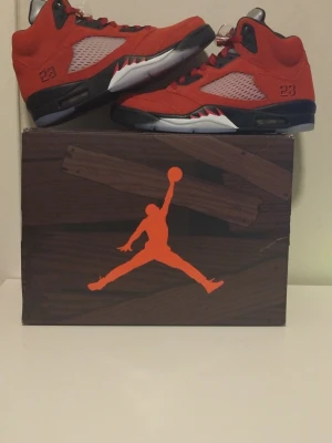 Nike Air Jordan 5 Röd Mocka - Säljer ett par Nike Air Jordan 5 i röd mocka med svarta och vita detaljer. Skorna har silvriga tungor, genomskinliga meshpaneler på sidorna och klassisk snörning. Sulan är svart och vit med isblå detaljer undertill. Ikoniska Jumpman-loggor på häl och tunga.