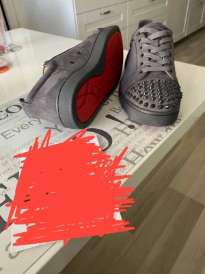 Grå mocka sneakers från Louboutin - Säljer ett par gråa sneakers från Christian Louboutin i mocka med ikonisk röd sula och coola nitar på tån. Skorna har snörning och en rund tå, vilket ger en edgy vibe. Perfekta för dig som vill sticka ut med exklusiva detaljer och lyxig känsla.