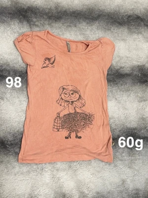 Aprikosfärgad t-shirt med tryck i storlek 98 - Söt aprikosfärgad t-shirt med korta ärmar och ett unikt tryck av en tecknad tjej med glasögon, kjol och fågelbur samt en liten fågel på bröstet. Perfekt för dig som gillar lekfulla motiv och mjuka material.🌸 Fint använt skick. Storlek 98
