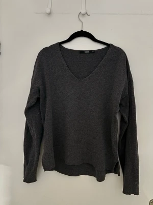 Mörkgrå stickad tröja från Bik Bok - Mysig mörkgrå stickad tröja från Bik Bok med v-ringad hals och långa ärmar. Tröjan har en avslappnad passform och ribbade muddar vid ärmslut och nederkant. Perfekt för lager på lager under kyliga dagar.