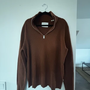 Brun ribbad stickad tröja med half zip - Säljer en brun ribbad stickad tröja med half zip och hög krage från Jack & Jones. Tröjan har lång ärm och en relaxed passform, perfekt för lager på lager under höst och vinter. Enkel men snygg design som funkar till det mesta.