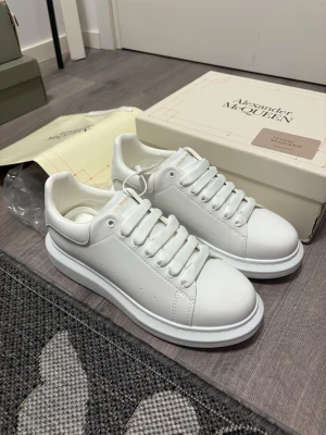 Vita sneakers från Alexander McQueen - Säljer ett par klassiska vita sneakers från Alexander McQueen med chunky plattformssula och guldfärgad logga på häl och plös. Skorna har rund tå, breda vita skosnören och är tillverkade i slätt skinn. Perfekta för dig som vill ha en clean och trendig look.