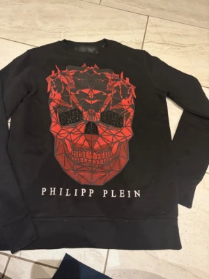 En Svart Philipp Plein sweatshirt med röd dödskalle - Cool svart sweatshirt från Philipp Plein med stor röd geometrisk dödskalle framtill och logga under. Tröjan har långa ärmar och ribbade muddar. Materialet känns mjukt och skönt, troligen bomull eller bomullsblandning. Perfekt för dig som gillar statement-plagg.