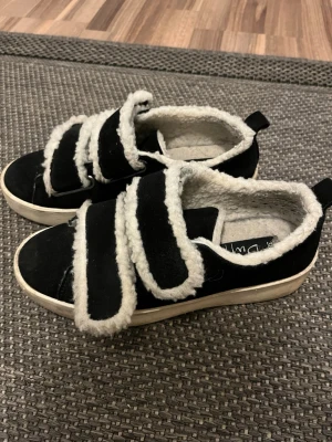 Svarta sneakers med fuskpäls från Duffy - Svarta sneakers från Duffy med tre breda kardborreband och vit fuskpäls längs kanterna och insidan. Skorna har en platt vit sula och rund tå. Perfekta för dig som vill ha både stil och mysfaktor under kyligare dagar.🤩🤩