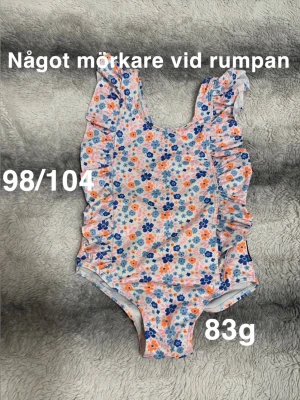 Blommig baddräkt med volanger - Söt baddräkt med blommigt mönster i blått, orange och rosa. Volangdetaljer längs axelbanden och sidorna ger en gullig touch. Perfekt för strand eller poolhäng.🌸 Inte använd många gånger men lite mörkare vid rumpan ( upplever inte att det syns på) storlek 98/104