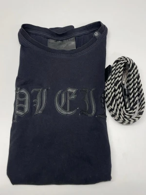 Svart t-shirt från Philipp Plein - Cool svart t-shirt från Philipp Plein med tryckt logga i gotisk stil på bröstet och en snygg läderdetalj med dödskalle vid nederkanten. Klassisk passform och rund halsringning. Perfekt för dig som gillar streetwear och exklusiva märken. Man får även med ett bälte. Skriv angående frågor!