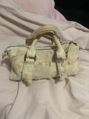 Beige handväska i sliten denim - Cool beige handväska i denim med slitna detaljer och grova sömmar. Väskan har två rejäla handtag och en stor dragkedja upptill. Perfekt för dig som gillar en edgy och avslappnad stil. Unik look som sticker ut!