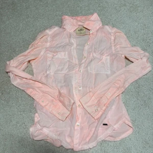 Ljusrosa skjorta från Hollister XS - Säljer en ljusrosa skjorta från Hollister i storlek XS. Skjortan har klassisk krage med spetsdetalj, två bröstfickor och knappar hela vägen fram. Tillverkad i tunn bomull, perfekt för lager på lager och chill stil.