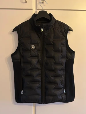 Svart dunväst från Spooks - Svart dunväst från Spooks med quiltad framsida och mjuk baksida. Västen har dragkedja framtill, två fickor med dragkedja och hög krage. På bröstet finns ett litet märke med texten 'Equestrian Sports' och på ryggen står det 'SPOOKS' i stora bokstäver.