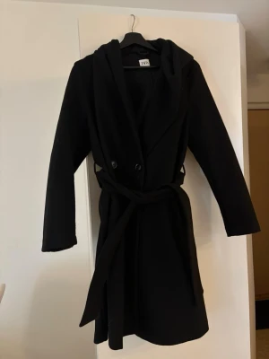 Svart kappa med huva från Zara - Svart kappa från Zara med huva och bälte i midjan. Klassisk och stilren look med knappar framtill och långa ärmar. Perfekt för dig som gillar enkel design och vill ha en jacka som håller dig varm under höst och vinter. För mig som är 165 cm går den strax under knäskålen. Fler bilder finns tyvärr inte då den ej passar längre. Köpt för tre år sen för 699, säljer för 300!