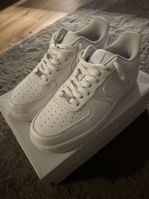 Nike Air Force 1 helvita sneakers - Nike Air Force 1 sneakers i helvit färg med klassisk låg siluett. Skorna har snygg snörning, perforerad tå och den ikoniska swoosh-loggan på sidan. Tillverkade i skinn med platt sula och rund tå, perfekt för dig som gillar cleana och stilrena sneakers.