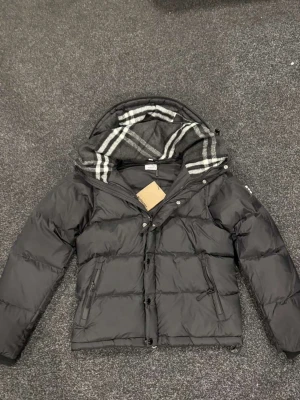 Svart pufferjacka från Burberry - Svart pufferjacka från Burberry med klassisk rutig huva i grått och vitt. Jackan har dragkedja och knappar framtill, två sidofickor med dragkedja och quiltad design. Perfekt för kalla dagar med sin tjocka och fluffiga look.