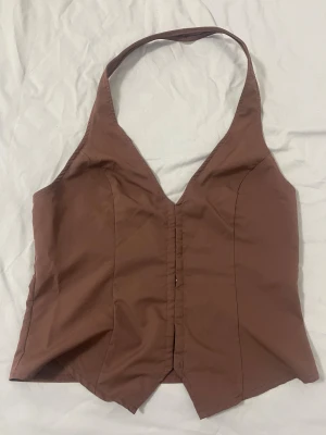 Brun korsett-topp från SHEIN - Snygg brun korsett-topp från SHEIN med halterneck och v-ringning. Toppen har en figurnära passform och är tillverkad i mjuk polyester. Framtill finns en markerad söm och spetsig nederkant som ger en cool look. Perfekt för dig som vill sticka ut lite extra.