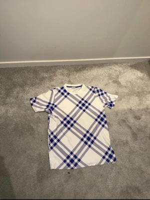 Vit rutig Burberry t-shirt - Säljer en snygg vit t-shirt från Burberry med blått och grått rutmönster. T-shirten har klassisk rund hals och korta ärmar. Materialet är mjuk bomull som känns skönt mot huden. Perfekt för dig som gillar stilrena designer med en twist.