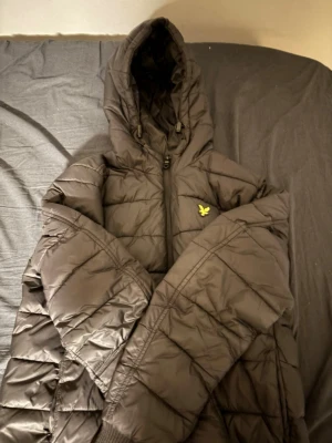 Svart pufferjacka från Lyle & Scott - Svart pufferjacka från Lyle & Scott med huva och gul logga på bröstet. Jackan har quiltad design, dragkedja framtill Perfekt för kalla dagar och har en clean, sportig look som funkar till streetwear.