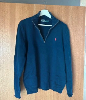 Marinblå stickad tröja Polo Ralph Lauren - Snygg marinblå stickad tröja från Polo Ralph Lauren i storlek S. Tröjan har half zip, ribbad krage och muddar samt den klassiska röda logon broderad på bröstet. Tillverkad i 100% bomull, perfekt för dig som gillar stilrena och tidlösa plagg.