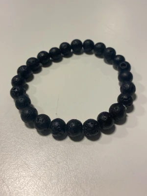 Svart pärlarmband med lava-look - Snyggt svart armband med runda pärlor som har en cool, lite grov lava-sten look. Elastiskt band gör det enkelt att ta på och av. Perfekt accessoar för dig som gillar minimalistisk stil med en twist.