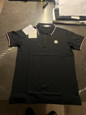 Svart Moncler piké med röd/vit kant - Står M men passar både S och M om man vill ha en tajtare look. Svart pikétröja från Moncler med klassisk krage och knappslå framtill. Röd och vit randig kant på krage och ärmslut ger en snygg kontrast. Moncler-logga broderad på bröstet. Tillverkad i mjuk bomull som känns skön mot huden.