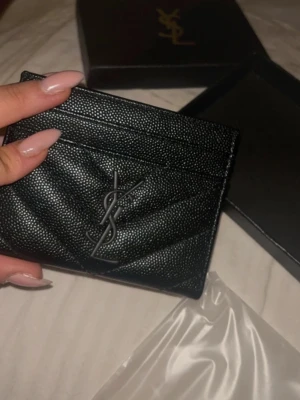 Svart YSL plånbok i skinn - Snygg svart plånbok från Saint Laurent med logo framtill. Gjord i strukturerat skinn med quiltad design och flera kortfack. Perfekt storlek för att ha med sig överallt. 