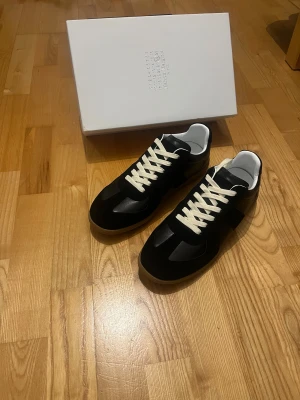 Maison Margiela sneakers i svart - Sneakers i svart med vita snören och gummisula. Klassisk low-top siluett med ovandel i skinn och mocka. Snygg minimalistisk design med diskreta detaljer och vit insida. Perfekta för dig som gillar stilrena och exklusiva sneakers.