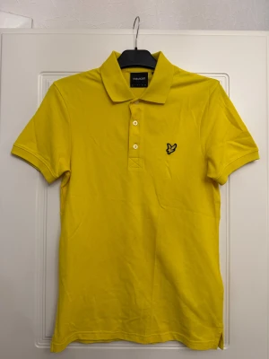 Gul piké från Lyle & Scott - Säljer en fräsch gul pikétröja från Lyle & Scott med klassisk krage och tre knappar framtill. Pikén har korta ärmar och den ikoniska fågel-loggan broderad på bröstet. Perfekt för dig som gillar färg och vill sticka ut lite extra.