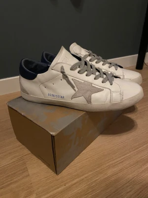 Golden Goose vita sneakers med stjärna - Golden Goose sneakers i vitt skinn med grå mockadetaljer och en stjärna på sidan. Skorna har grå snörning, mörkblå häl och svart platt sula. Snygg och clean design med coola kontraster och GGDB/SSTAR-tryck på sidan. Det finns en liten defekt på undersidan men inget man tänker på. 