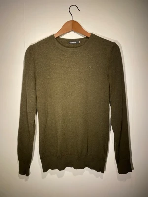 Olivgrön merinoulltröja J.Lindeberg - Snygg olivgrön tröja från J.Lindeberg i 100% merinoull. Klassisk rund hals och ribbade muddar vid ärmslut och nederkant. Liten broderad logga på bröstet. Perfekt för dig som gillar stilrena och sköna plagg med premium känsla. På lappen står det L men den är mer som S för att den krympte lite i tvätten