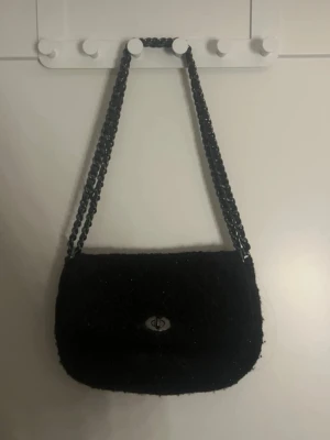 Svart axelväska med kedja - Snygg svart axelväska med fluffigt tyg och grov kedja som axelrem. Väskan har en rundad form och ett metallspänne framtill. Perfekt för dig som vill ha en stilren men ändå cool accessoar till din outfit. Väskan är från Beck södergarner🥰nypris 950, kan sälja för 339