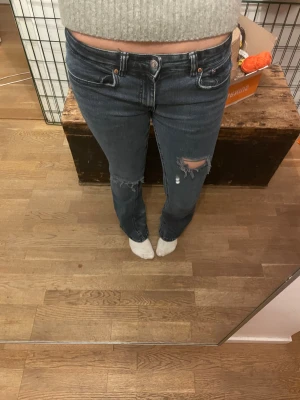 Mörkblå lågmidjade bootcut jeans med slitningar - Säljer ett par lågmidjade mörkblå jeans med bootcut passform och snygga slitningar på knä och benslut. Jeansen har klassisk femficksdesign, låg midja och är tillverkade i jeansmaterial. Perfekta för dig som gillar en avslappnad och trendig look.