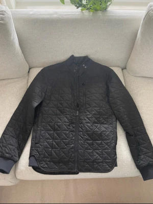 Svart quiltad jacka från Barbour - Quiltad svart jacka från Barbour International med snygg ståkrage och dragkedja framtill. Jackan har ett diskret Barbour-märke, ribbade muddar och ett stilrent rutmönster över hela ytan. Perfekt för dig som gillar klassisk och clean stil.